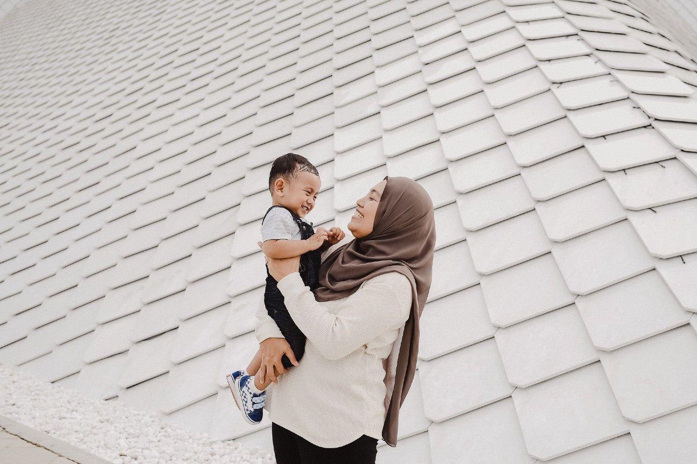 Mother in head scarf holding and smiling at toddler son.