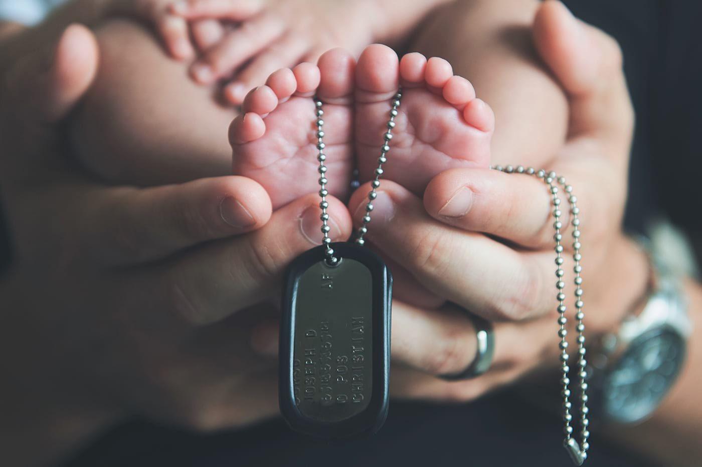 A parent cradles their baby's feet, which are draped with military tags.